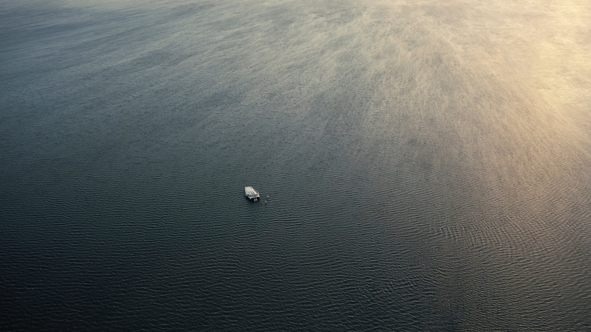 A tiny white shape floats alone on a vast expanse of dark, rippled water, seen from above