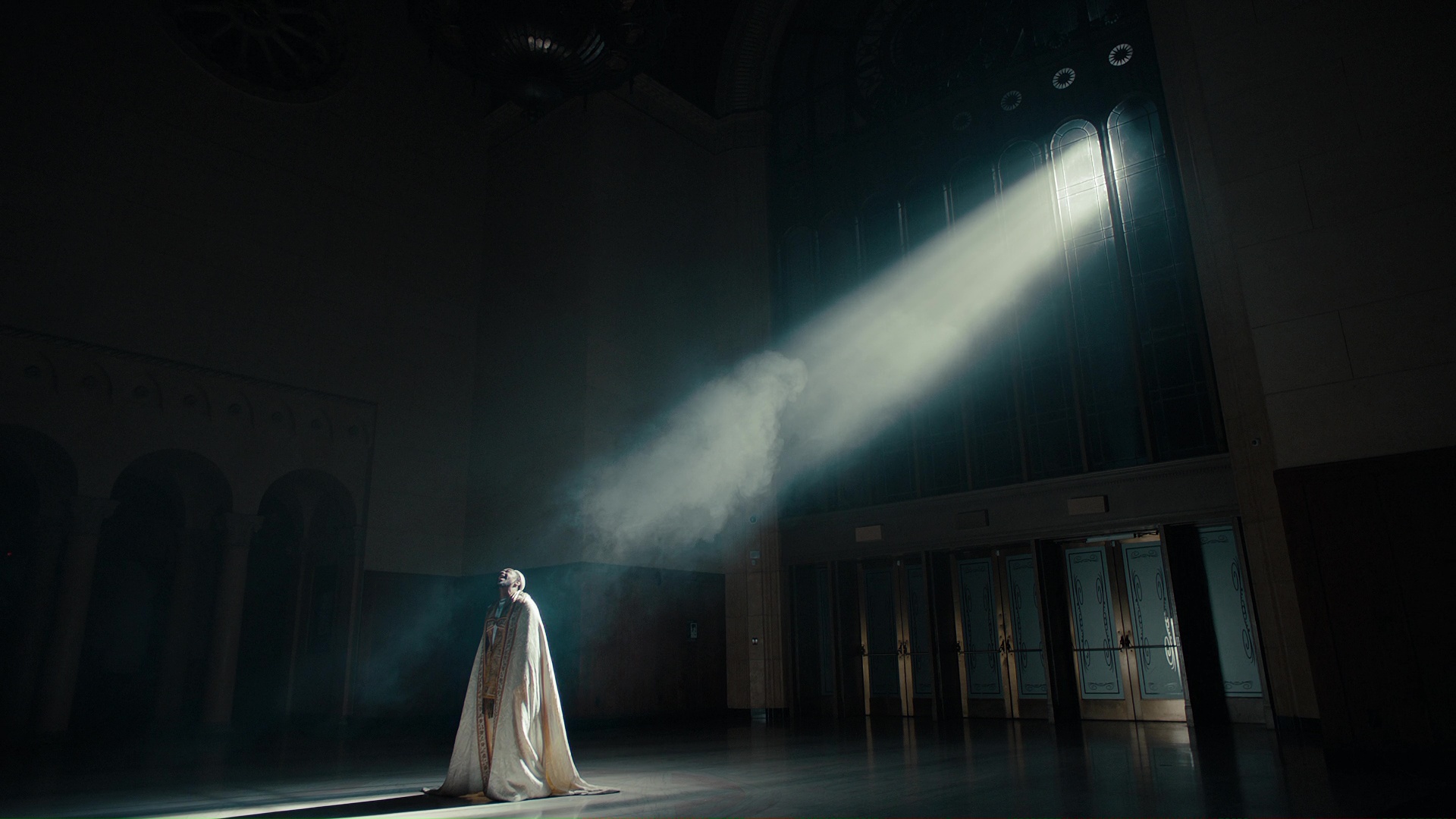 A figure in a pale robe stands at the foot of a diagonal shaft of light in a dark hall