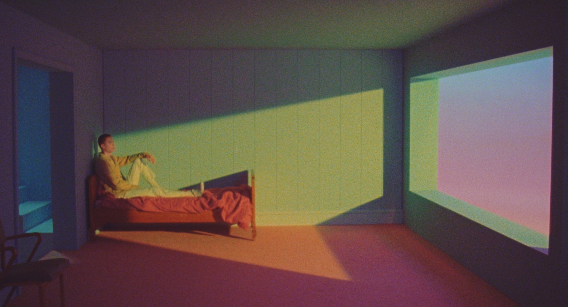 An animated figure sits on a bed in a room where bands of coloured light fall across the walls and floor from a large window