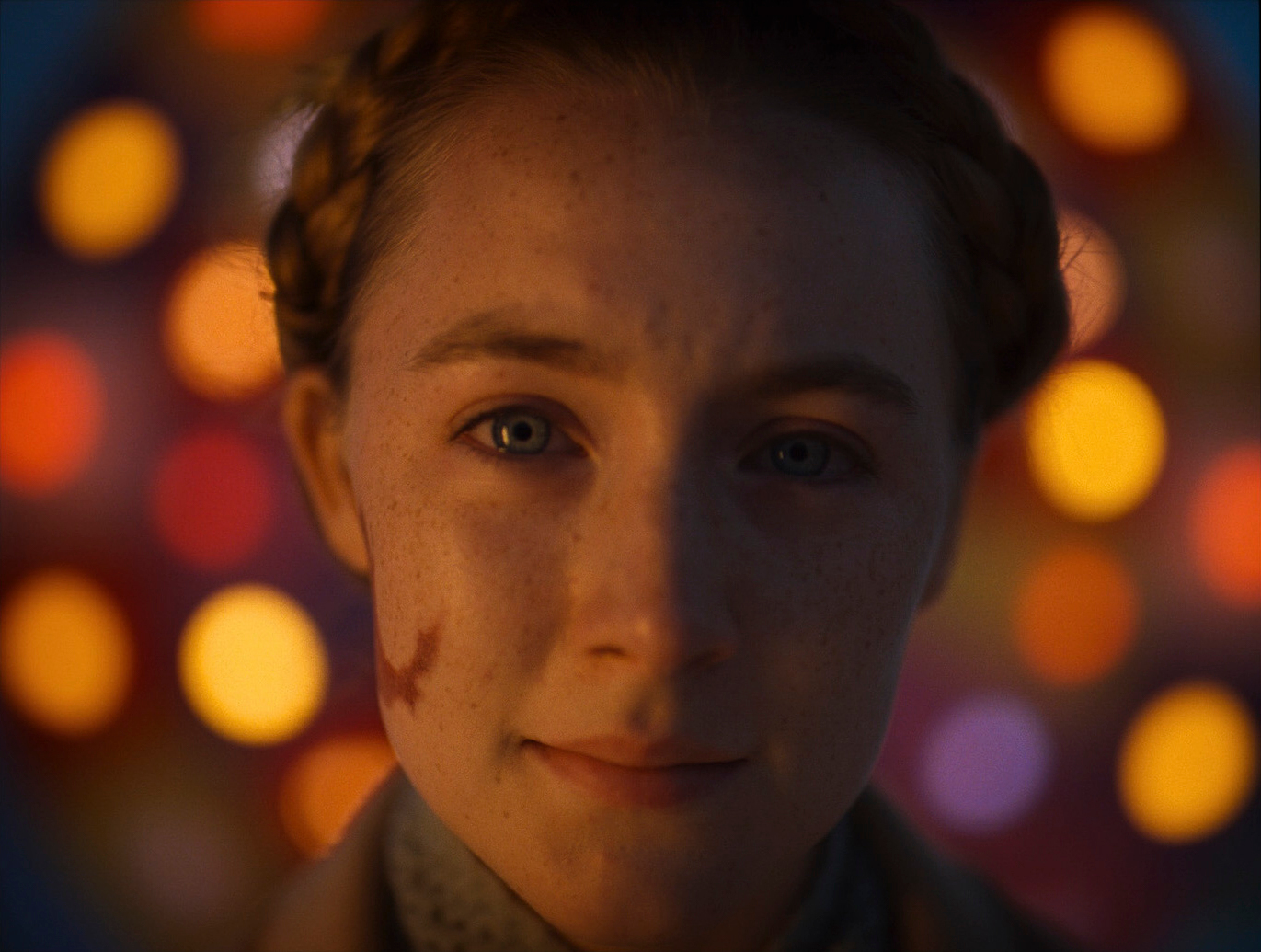 A close-up of a young woman's freckled face and plaited hair against soft orange out-of-focus lights