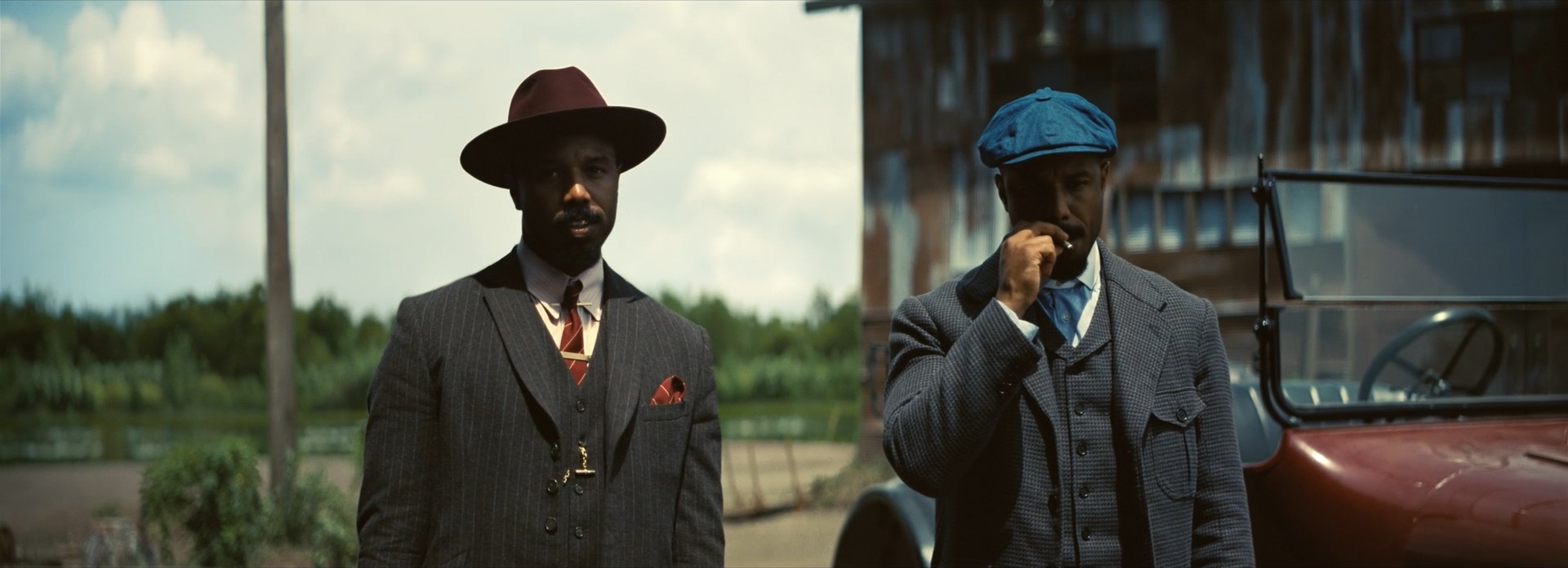 Two men in suits, one in a wide-brimmed hat and one in a flat cap, stand side by side beside a red vintage car in a green landscape