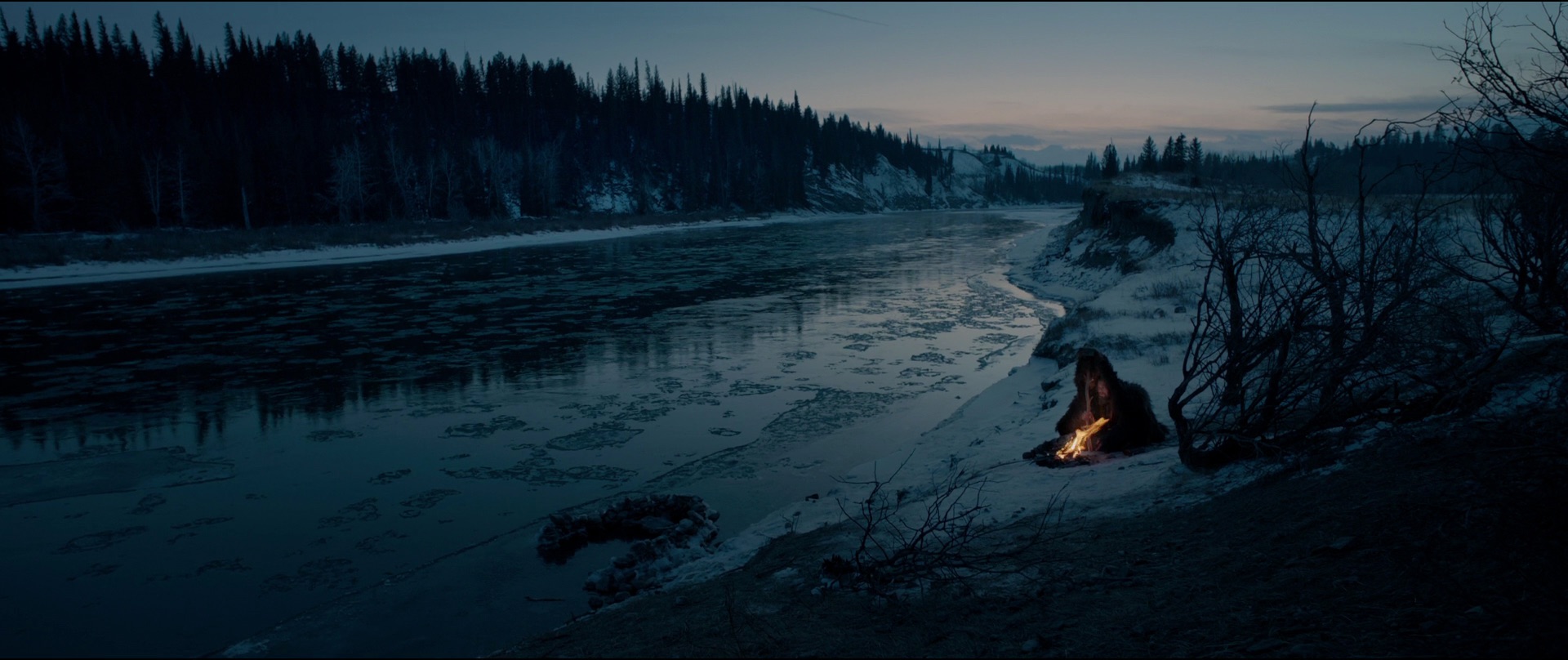 A wide dusk shot of a frozen river and pine forest, with a tiny figure crouched at a small fire on the bank