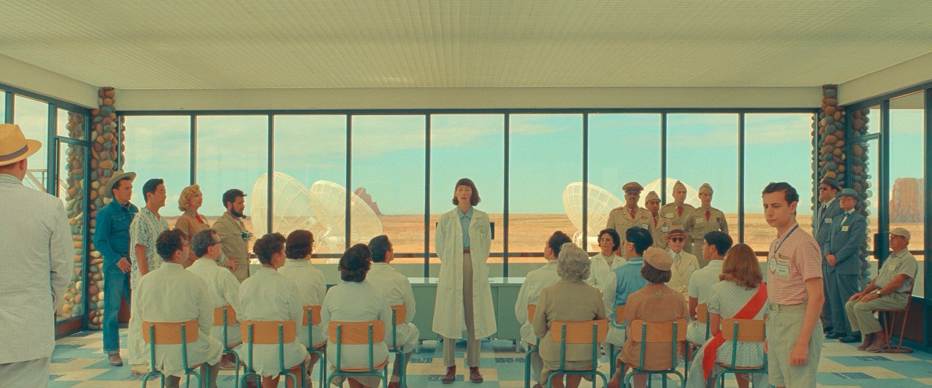 A symmetrical wide shot of a pale-blue room with tall windows, rows of people in white seated facing one person standing at the centre